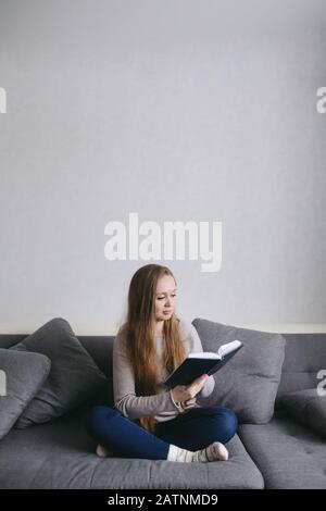 Intelligente junge Frau, die zu Hause auf der Couch sitzt und Buch liest. Schöne Frau mit langen Haaren im modernen minimalistischen Interieur. Hochformat in voller Länge, Co Stockfoto