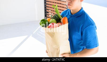 Mann für Lebensmittelzustellung in blauer Uniform. Thailändischer Lieferer, der frische Lebensmittel für junge Frau hält. Kunde erhält Auftrag vom Kurier zu Hause. Stockfoto