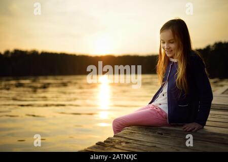 Süßes kleines Mädchen, das an einem warmen Sommertag auf einer Holzplattform am Fluss oder am See sitzt und ihre Füße im Wasser taucht. Familienaktivitäten im Sommer. Stockfoto