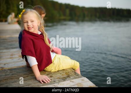 Zwei süße kleine Mädchen sitzen auf einer hölzernen Plattform durch den Fluss oder See Ihre Füße eintauchen in das Wasser an warmen Sommertagen. Aktivitäten für die ganze Familie in Summe Stockfoto