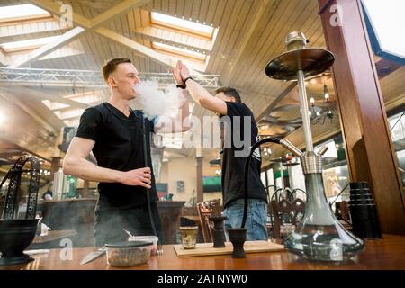 Hookah in der Prozessvorbereitung in der gemütlichen Hookah-Bar. Zwei junge Männer Shisha-Meister geben fünf, während sie Hookah für das Rauchen vorbereiten. Stockfoto