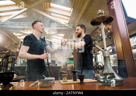 Hookah in der Prozessvorbereitung in der gemütlichen Hookah-Bar. Zwei junge Männer Shisha-Meister geben fünf, während sie Hookah für das Rauchen vorbereiten. Stockfoto