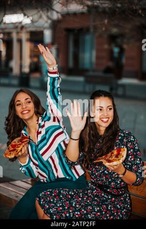 Zwei schöne junge Frauen, die im Freien Pizza essen und ihre Freunde winken. Lifestyle, modernes Leben. Stockfoto