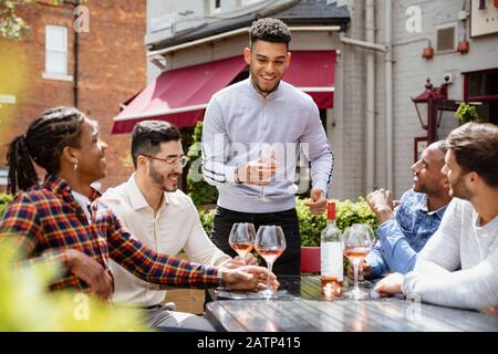 Eine Gruppe von männlichen Freunden, die zusammen ein Glas Wein im Freien im Biergarten der Stadt genießen. Einer von ihnen hält eine Festrede. Stockfoto