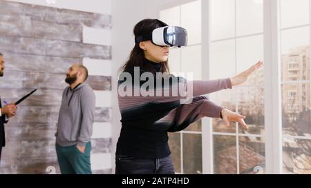 Architektin verwendet virtuelle Reality-Brille in leerer Wohnung und Immobilienmakler, die im Hintergrund mit dem Kunden sprechen. Stockfoto