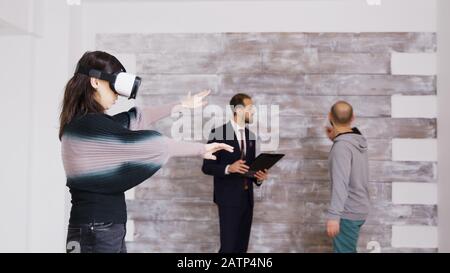 Designerin mit virtueller Reality-Brille in leerer Wohnung, während Immobilienmakler im Hintergrund mit dem Hausbesitzer spricht. Stockfoto