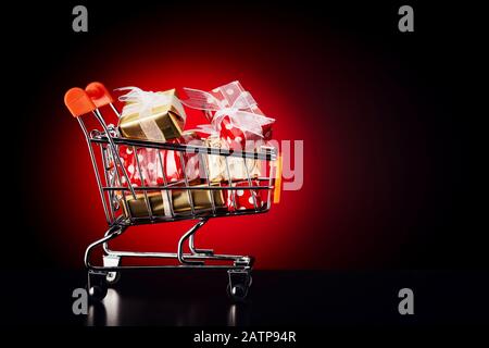 Einkaufswagen mit bunten Geschenkschachteln auf rotem Hintergrund. Kaufkonzept für weihnachten oder den schwarzen freitag. Stockfoto
