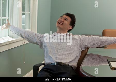 Junger asiatischer Geschäftsmann sitzt im Stuhl am Arbeitsplatz und feiert Ein Gewinn Stockfoto