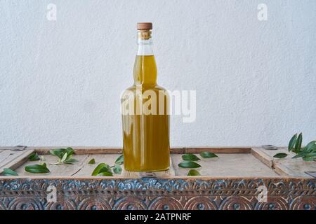 Stillleben einer Glasflasche auf einem rustikalen Holzmöbel, gefüllt mit frischem Olivenöl, umgeben von grünen Olivenblättern an einer weißen Wand Stockfoto