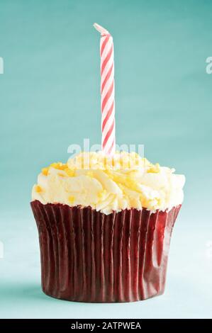 Ein einziger Geburtstagskuchen mit einer gestreiften roten und weißen Kerze in brauner Papierkoffer mit gelber Buttercremefarbe und bestreuten. Türkisfarbener Hintergrund. Stockfoto