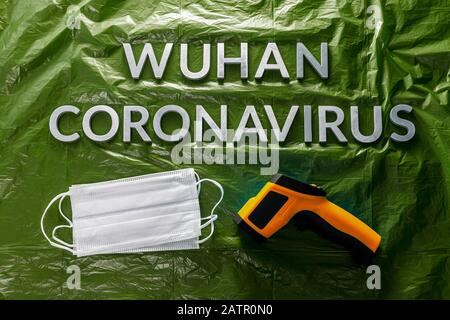 Worte wuhan Coronavirus mit Metallbuchstaben auf grün zerknitterter Kunststoff-Filmkulisse mit Gesichtsmasken und Infrarotthermometer - flach lag mit Drama Stockfoto