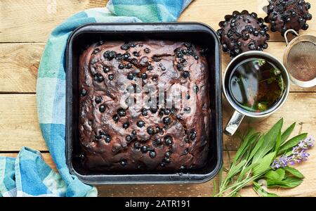 Schokoladenbeerkuchen mit Cupcakes und Kräutern: Minze, Lavendel, Salbei in der Nähe und Tasse Thymiantee auf rustikalem Holztisch, flacher Lay, von oben nach oben, Stockfoto