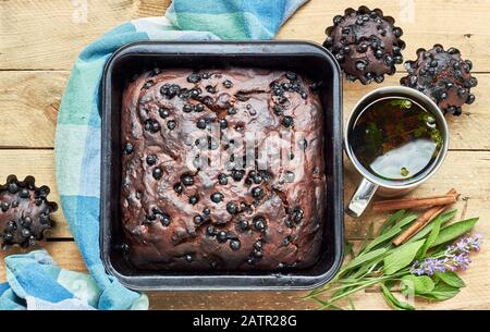 Schokoladen-Blaubeerkuchen mit Cupcakes und Kräutern: Minze, Lavendel, Salbei und Zimt in der Nähe und Tasse Thymian-Tee auf rustikalem Holztisch, flacher Lay, Fro Stockfoto