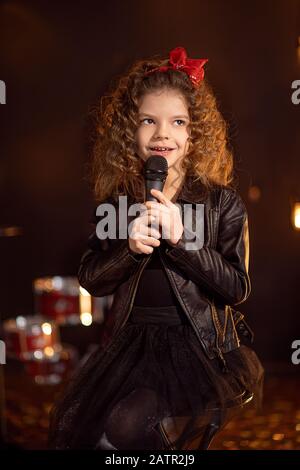 Schönes Mädchen mit lockigen Haaren, Lederjacke, Stiefel singen in ein drahtloses Mikrofon für Karaoke wie Rockstar im Tonstudio oder auf der Bühne. Stockfoto