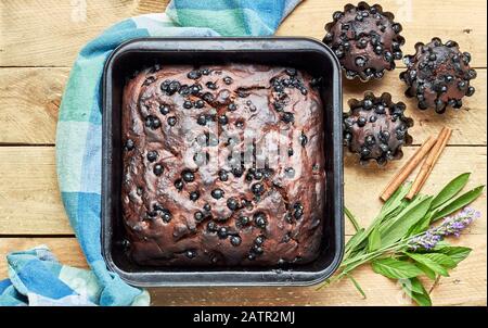 Schokoladenbeerkuchen mit Cupcakes und Kräutern: Minze, Lavendel und Salbei und Zimt in der Nähe auf rustikalem Holztisch, flacher Lay, von oben nach oben, nah Stockfoto