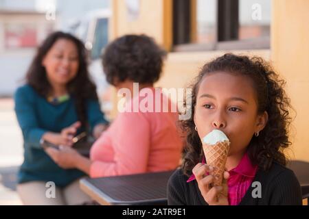 Junges Mädchen genießt ein Eis Kegel wie ihre Mutter Und Großmutter sitzt hinter ihr reden Stockfoto