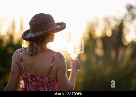 Junge Frau, die einen Sonnenhut trägt und ein Glas Weißwein draußen in der untergehenden Sonne hält. Stockfoto