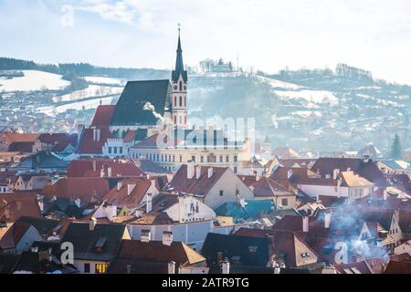 Blick auf den Sonnenaufgang auf die Kirche Cesky Krumlov Saint Vitus im Winter und die kleine Stadt um sie herum. Schneebedeckte Dächer mit Raucherkanimsen. Stockfoto