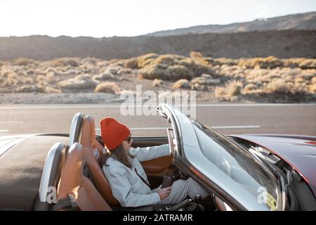 Frau, die während eines Sonnenuntergangs auf der Wüstenstraße ein Cabriolet-Auto fährt. Sorgloses Lifestyle- und Reisekonzept