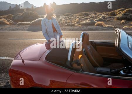 Frau, die wunderschöne felsige Landschaften genießt, während sie mit einem Auto während eines Sonnenuntergangs am Straßenrand der Wüste steht. Reisen im vulkanischen Tal auf Teneriffa Stockfoto