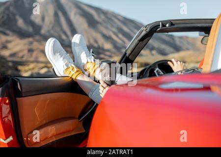 Frau, die mit einem Cabriolet im Wüstental unterwegs ist und die Beine am Straßenrand aus dem Autofenster zieht. Nahaufnahme auf weißen Sneakers und Berg im Hintergrund Stockfoto