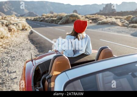 Frau, die schöne felsige Landschaften genießt, sitzt auf der Autohaube am Straßenrand während eines Sonnenuntergangs. Reisen im vulkanischen Tal auf Teneriffa Stockfoto