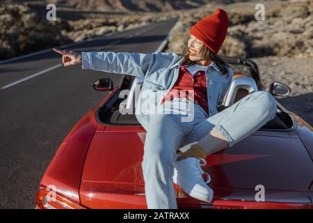 Frau, die schöne felsige Landschaften genießt, sitzt auf der Autohaube am Straßenrand während eines Sonnenuntergangs. Reisen im vulkanischen Tal auf Teneriffa Stockfoto