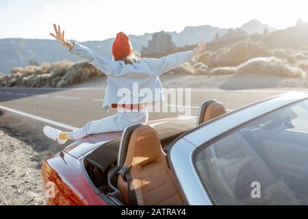 Frau, die schöne felsige Landschaften genießt, sitzt auf der Autohaube am Straßenrand während eines Sonnenuntergangs. Reisen im vulkanischen Tal auf Teneriffa Stockfoto