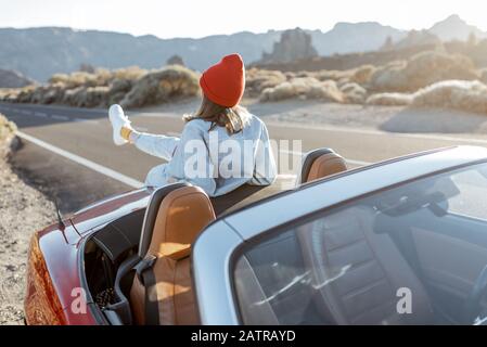 Frau, die schöne felsige Landschaften genießt, sitzt auf der Autohaube am Straßenrand während eines Sonnenuntergangs. Reisen im vulkanischen Tal auf Teneriffa Stockfoto