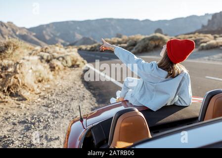 Frau, die schöne felsige Landschaften genießt, sitzt auf der Autohaube am Straßenrand während eines Sonnenuntergangs. Reisen im vulkanischen Tal auf Teneriffa Stockfoto