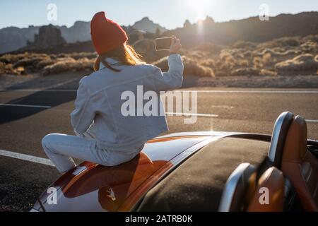 Frau, die schöne felsige Landschaften genießt, sitzt auf der Autohaube am Straßenrand während eines Sonnenuntergangs. Reisen im vulkanischen Tal auf Teneriffa Stockfoto