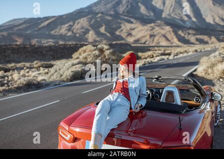 Frau, die schöne felsige Landschaften genießt, sitzt auf der Autohaube am Straßenrand während eines Sonnenuntergangs. Reisen im vulkanischen Tal auf Teneriffa Stockfoto