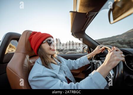 Fröhliche Frau in rotem Hut, die während eines Sonnenuntergangs auf der Wüstenstraße mit wunderschöner Felslandschaft im Hintergrund ein Cabriolet-Auto fährt Stockfoto