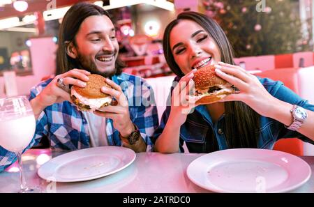 Ein Paar liebt es, zusammen Spaß zu haben, Burger im Café zu essen und zu lachen. Schmackhafter Milchshake im Weinglas, weiße Teller. Breites Lächeln, Make-up im Gesicht Stockfoto