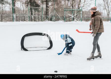 Kleiner lustiger Junge mit ihrer Mutter, die im Park Schlittschuhlaufen macht. Spielen Sie Eishockey mit Stick und Tor. Im Freien. Wintersport Stockfoto