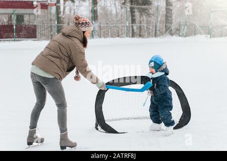 Kleiner lustiger Junge mit ihrer Mutter, die im Park Schlittschuhlaufen macht. Spielen Sie Eishockey mit Stick und Tor. Im Freien. Wintersport Stockfoto
