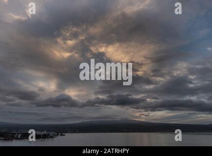 Hilo, Hawaii, USA. - 14. Januar 2020: Spektakuläre dunkle Abendwolkenlandschaft mit gelben Sonnenflecken über Stadt und Vulkan am Horizont. Stockfoto