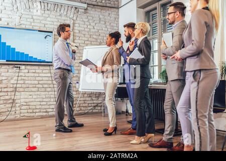 Respektabler älterer Mann in einem Business-Anzug, der in seinem Büro Minigolf spielt. In der Nähe sind seine Assistenten und das Zuschauen Stockfoto