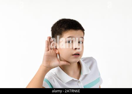 Fröhlicher kleiner Junge im gestreiften T-Shirt, der auf weißem Hintergrund eine Hörgeste macht. Gesichtsausdruck. Kind mit Hand über Ohr Hör- und Tratsch. Stockfoto