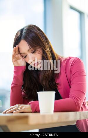 Eine professionelle asiatische Geschäftsfrau, die sich völlig überwältigt und traurig fühlt, während sie eine Pause in einem Café an ihrem... Stockfoto
