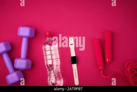 Smart Watch, Flasche Wasser, rotes Springseil und Flieder Hanteln auf rotem Hintergrund von einer Matte für Yoga, Draufsicht, Sportkulisse Stockfoto