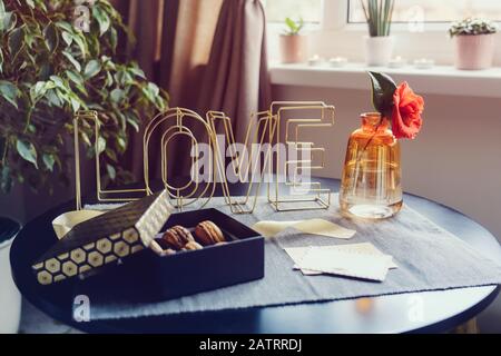 Festliche Schachtel mit Schokoladenmakaroon-Kuchen, Rosenblume in Vase, Postkarte und Drahtwort Liebe auf dem schwarzen Tisch in der Nähe des Fensters. Gemütlicher Valentinstag. Rom Stockfoto