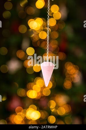 Rosafarbenes Liebes-Rosenquarz-Naturkristallpendel, das in Innenräumen an der Silberkette hängt, mit runden gelben Bokeh-Lichtern im Hintergrund. Vorhersage für neue Jahre Stockfoto