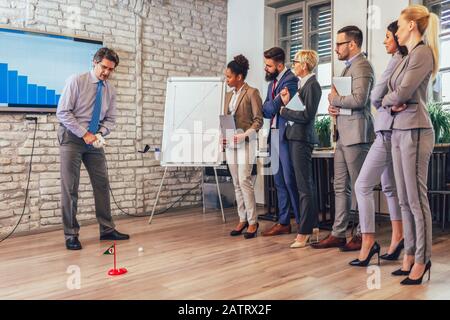 Respektabler älterer Mann in einem Business-Anzug, der in seinem Büro Minigolf spielt. In der Nähe sind seine Assistenten und das Zuschauen Stockfoto