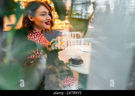 Blick Von Draußen Auf Die Schöne Brunette-Frau, Die Den Valentinstag Allein Mit Cupcake Im Gemütlichen Café Feiert. Konzept Der Selbstzufriedenheit Und Liebe T Stockfoto