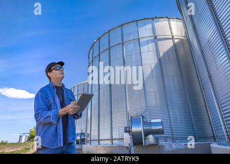 Ein Landwirt steht mit einem Tablet, während er neben steht und nach oben zu Getreidespeicher schaut; Alberta, Kanada Stockfoto