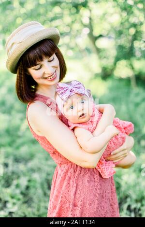 Schöne Mutter mit Strohhut Und ihrer kleinen Tochter im Freien sehen in einem rosafarbenen Kleid aus. Familienlook. Junge Mutter wirft Baby in den Stockfoto