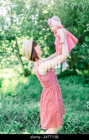 Schöne Mutter mit Strohhut Und ihrer kleinen Tochter im Freien sehen in einem rosafarbenen Kleid aus. Familienlook. Junge Mutter wirft Baby in den Stockfoto