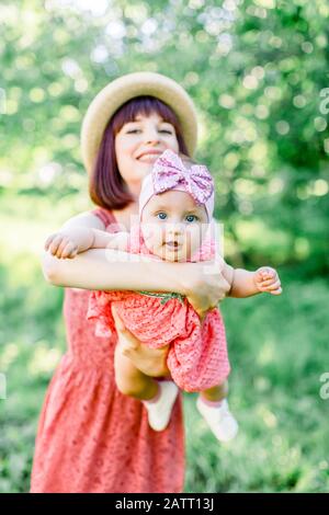 Schöne Mutter mit Strohhut Und ihrer kleinen Tochter im Freien sehen in einem rosafarbenen Kleid aus. Familienlook. Junge Mutter wirft Baby in den Stockfoto