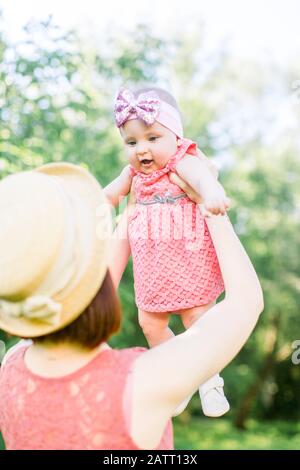 Schöne Mutter mit Strohhut Und ihrer kleinen Tochter im Freien, Familie, sehen in einem pinkfarbenen Kleid aus. Junge Mutter wirft Baby in den Himmel, auf sonnige Stockfoto